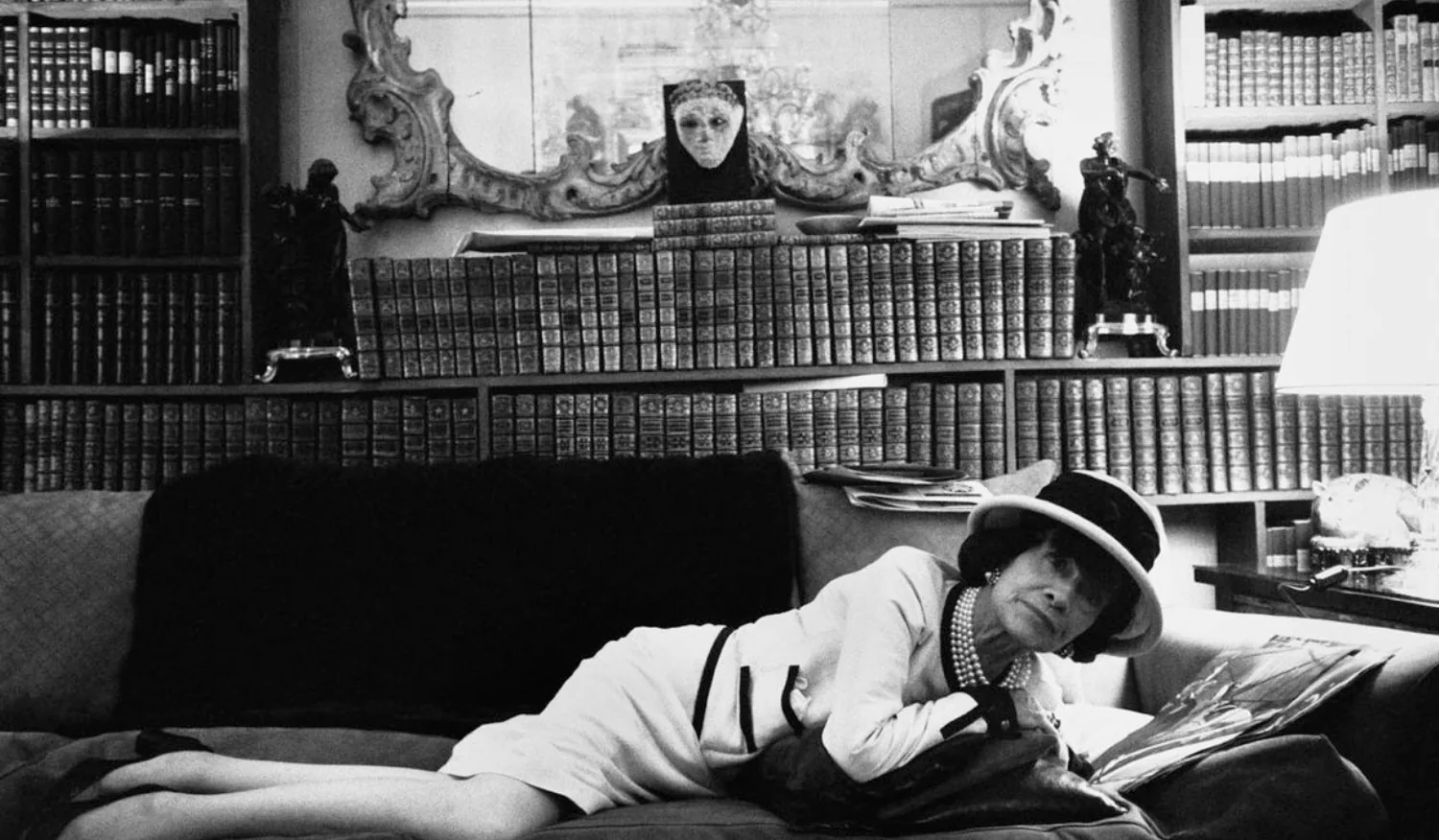 Коко Шанель в шляпе и светлом костюме лежит на диване на фоне книжных полок, чёрно-белая фотография